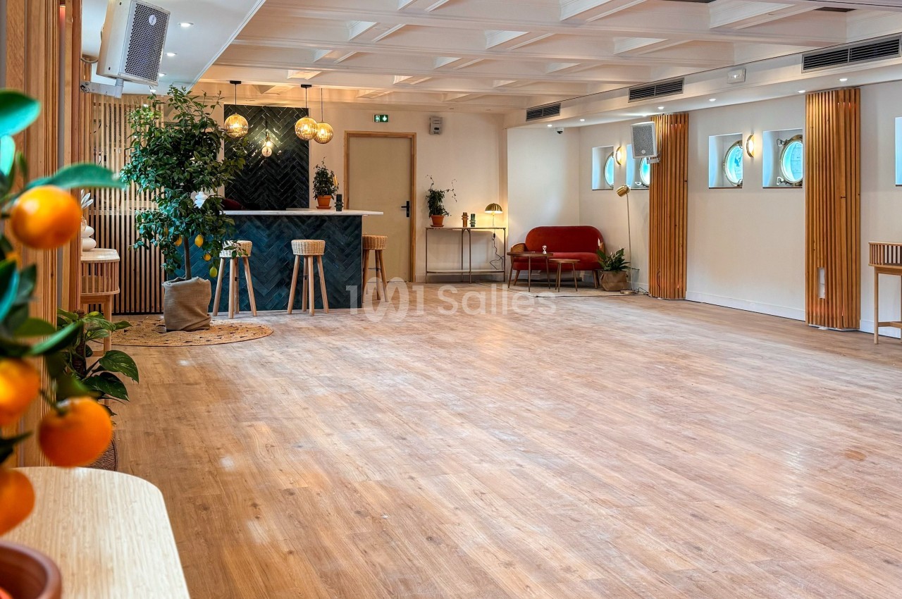 Salle lumineuse avec parquet en bois, bar au fond, plantes décoratives et mobilier moderne.