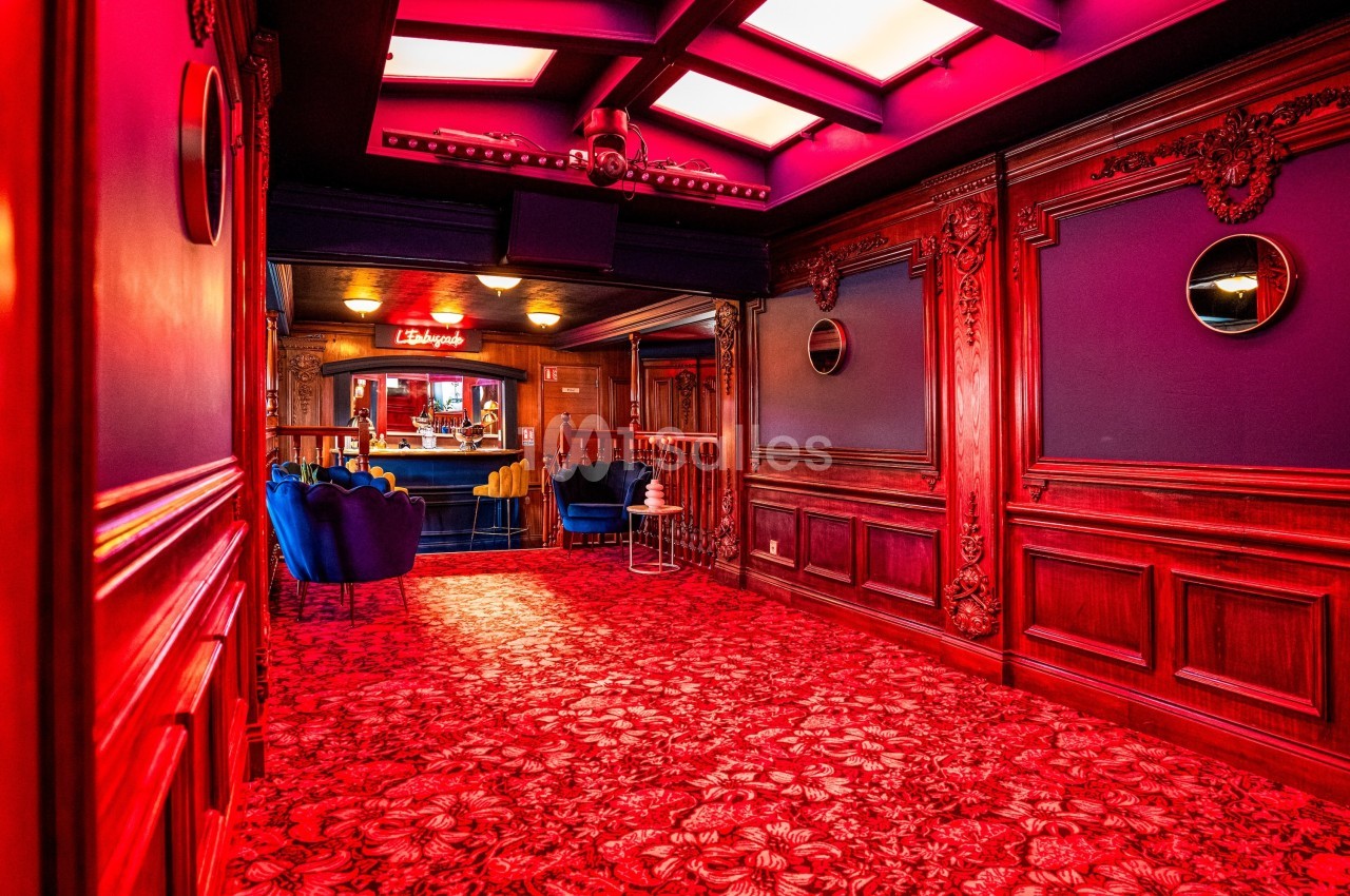 Couloir richement décoré avec tapis rouge à motifs, boiseries sculptées, et vue sur un espace lounge éclairé.