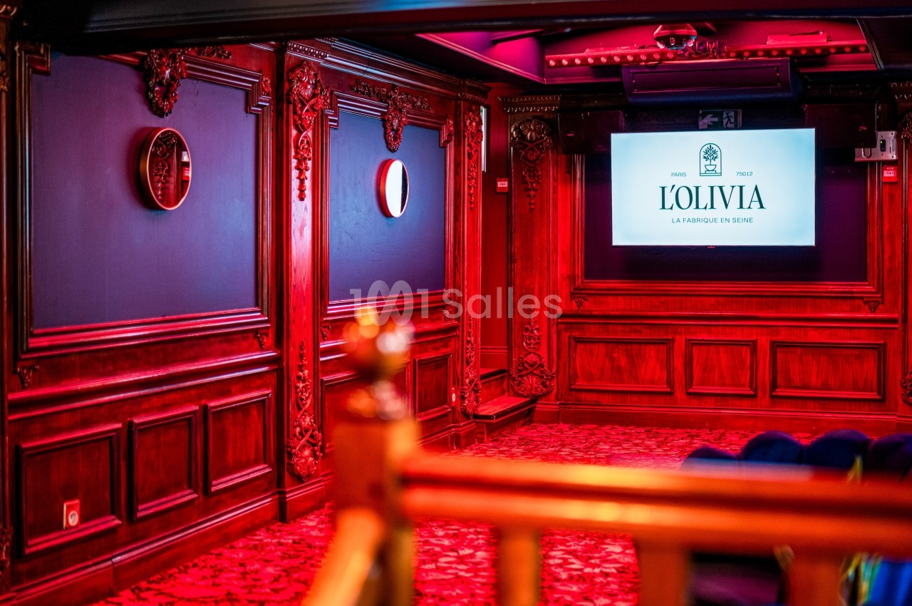 Salle décorée de boiseries rouges avec écran affichant ’L'Olivia’, sièges en bas et miroirs muraux.