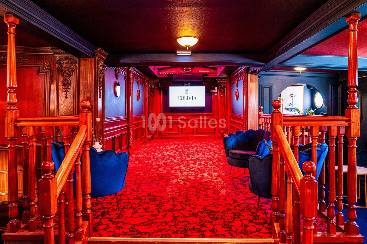 Salle élégante avec tapis rouge, fauteuils bleus, boiseries sculptées et écran affichant un logo au fond.