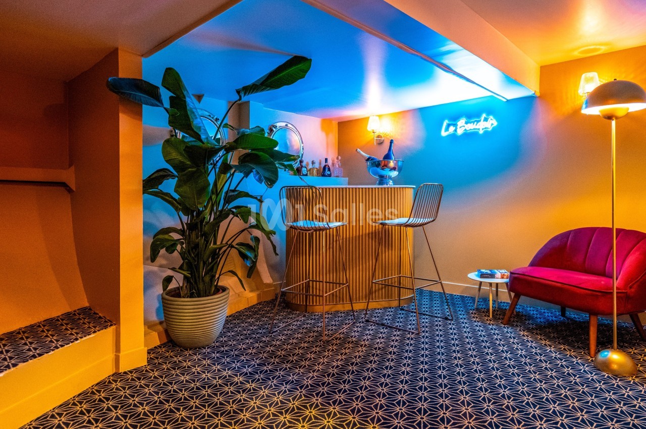 Coin bar intérieur avec tabourets, plante en pot, fauteuil rouge, éclairage chaleureux et néon bleu au mur.