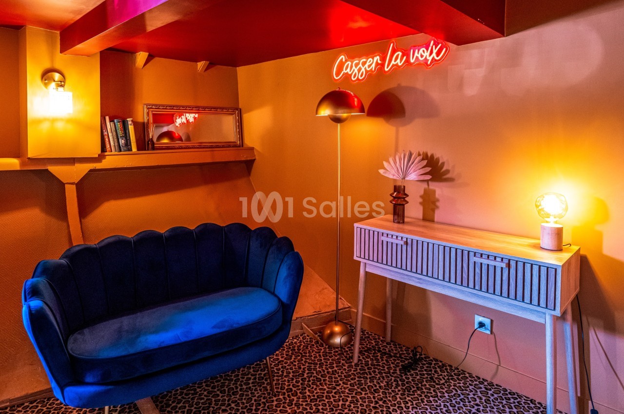 Salon cosy avec canapé bleu, lampe dorée, console blanche, miroir et néon rouge indiquant ’Casser la voix’.