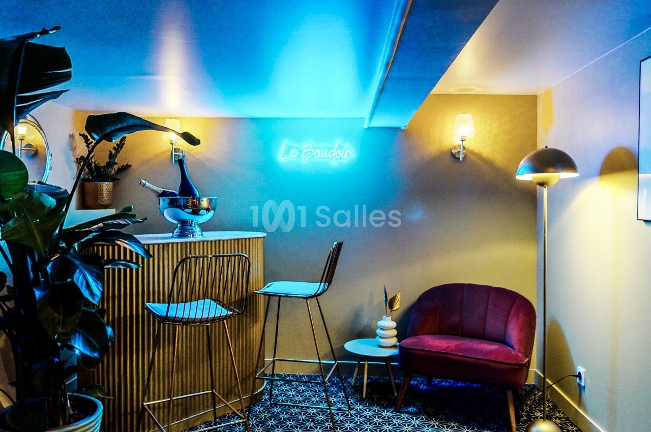 Coin salon avec fauteuil rose, bar en bois, plantes et éclairage néon bleu dans une ambiance tamisée.