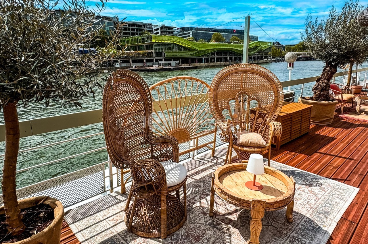 Mobilier en rotin disposé sur une terrasse en bois au bord d'une rivière, avec vue sur un bâtiment moderne et un pont.