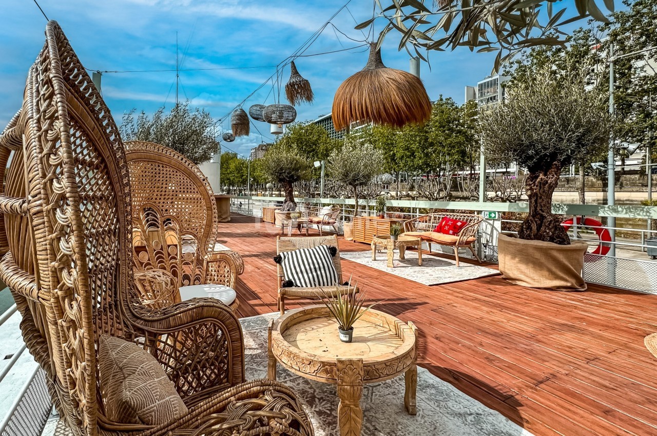 Terrasse en bois aménagée avec des fauteuils en rotin, des tables basses et des plantes, bordée par un cours d'eau.