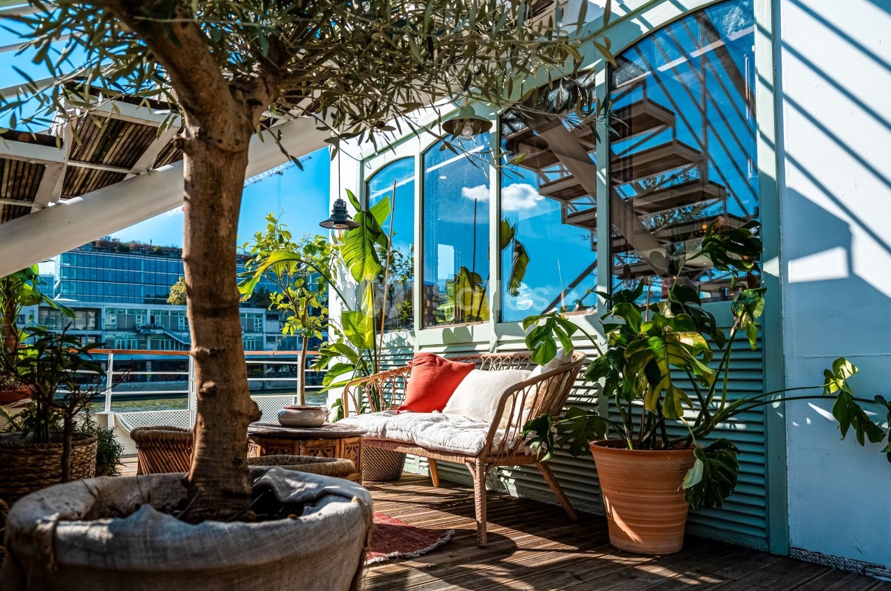 Terrasse ensoleillée avec canapé en rotin, coussins, plantes en pot et vue sur des bâtiments modernes au bord de l'eau.
