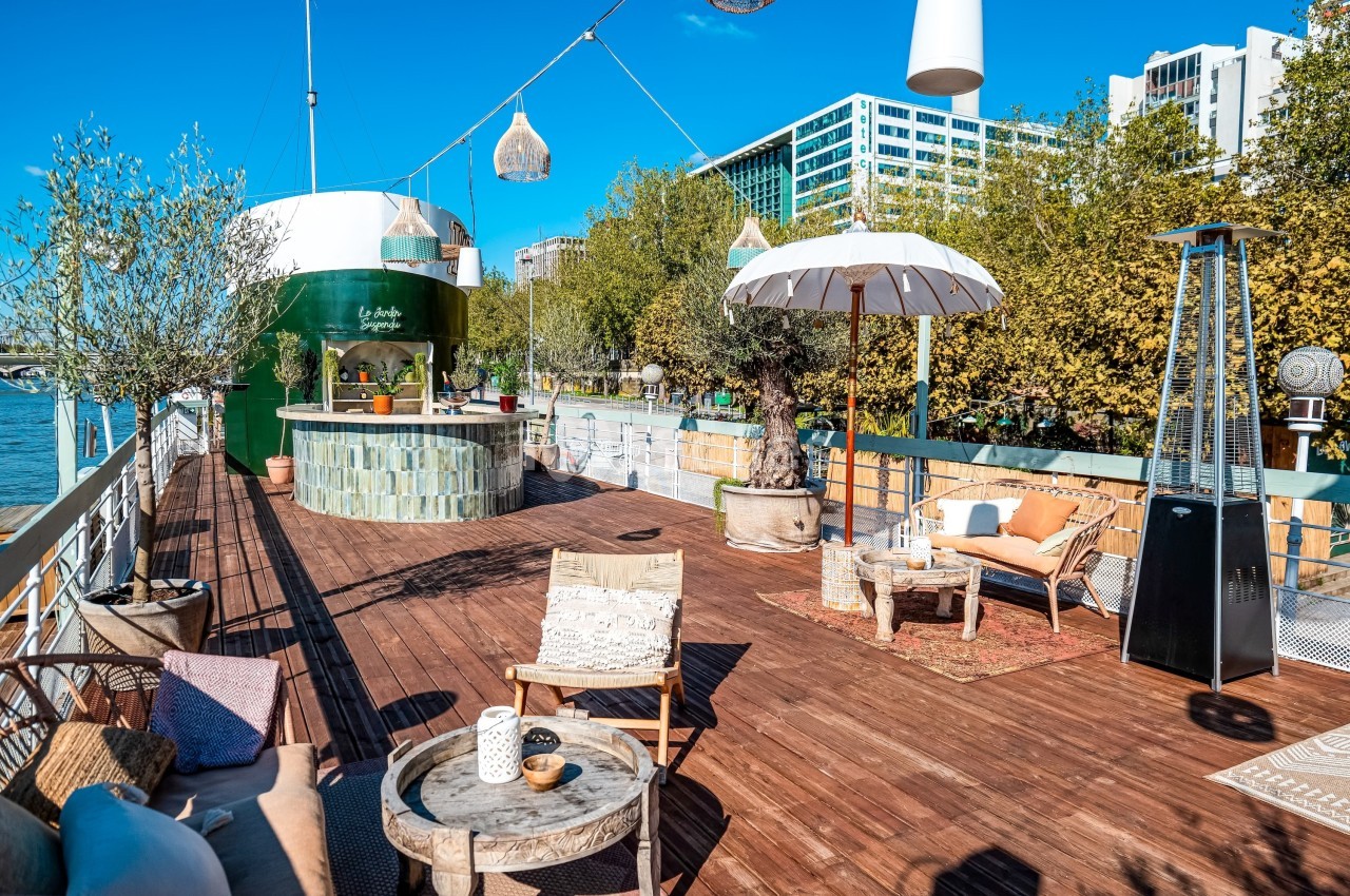 Terrasse en bois aménagée avec des fauteuils, parasols et plantes, située au bord d’un cours d’eau sous un ciel bleu.