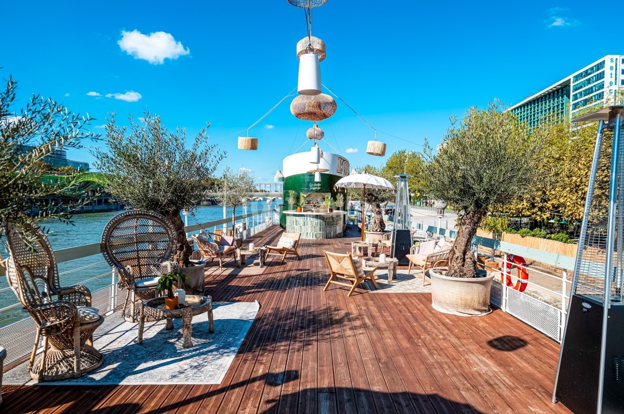 Terrasse en bois aménagée avec des fauteuils, des plantes en pot et une vue sur une rivière par temps ensoleillé.
