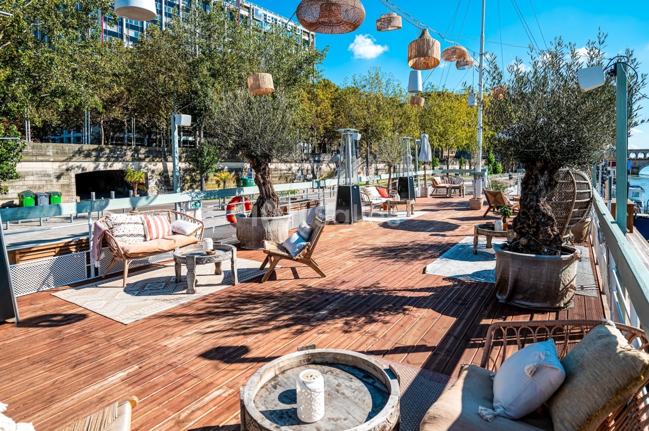 Terrasse en bois aménagée avec des fauteuils, des tables, des plantes et des lampes suspendues, sous un ciel ensoleillé.