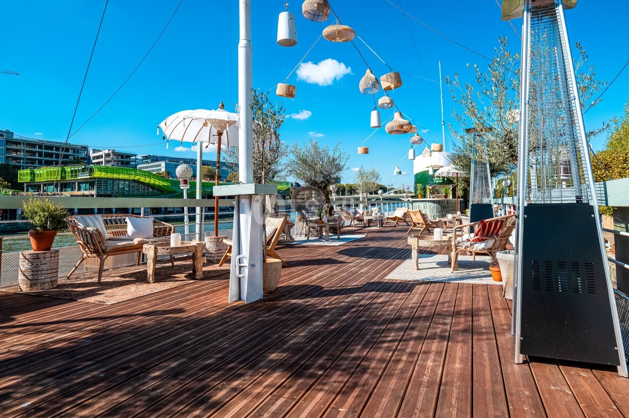 Terrasse en bois aménagée avec des fauteuils, parasols et suspensions, bordant un cours d'eau sous un ciel bleu.