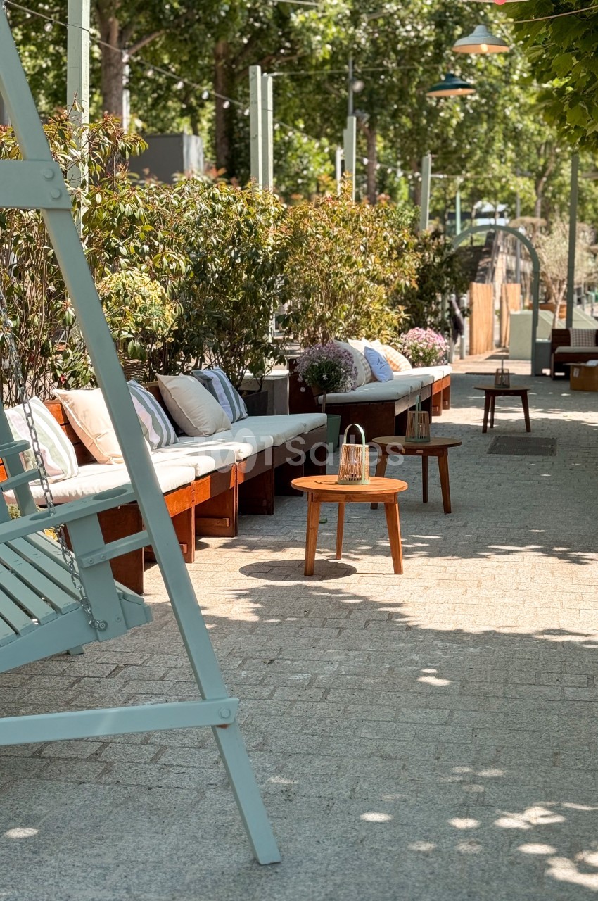 Terrasse extérieure avec bancs en bois, coussins, table basse et végétation sous un ciel ensoleillé.
