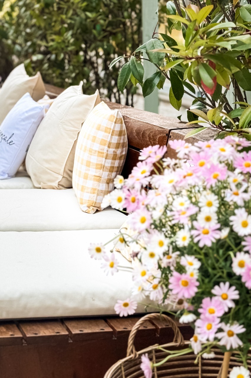 Banc en bois avec coussins beiges et à carreaux, entouré de verdure et de fleurs roses et blanches.