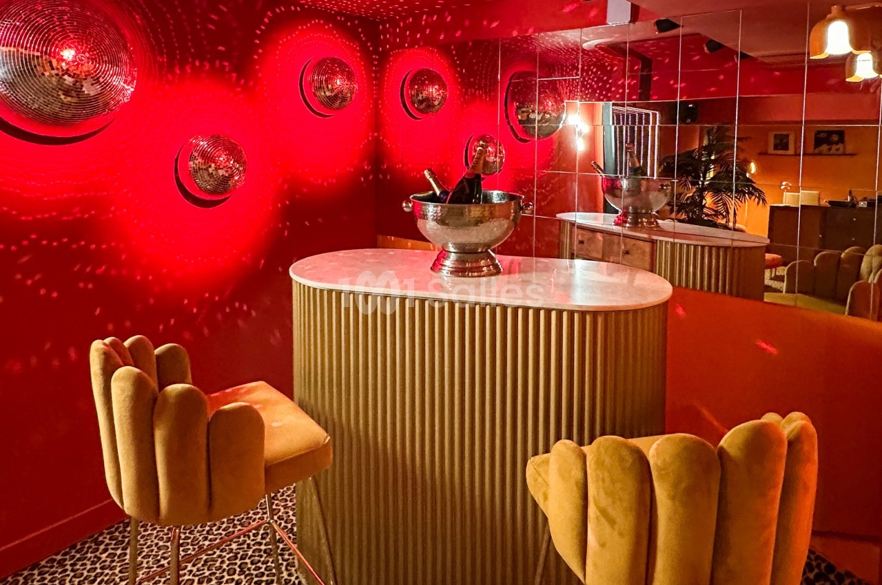 Bar intimiste avec murs rouges, table ovale en marbre, tabourets jaunes et éclairage tamisé reflété par des miroirs.