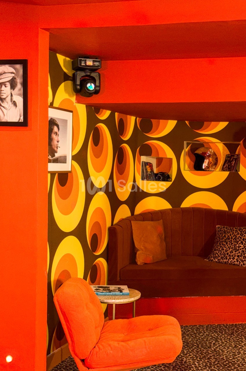 Salon rétro avec fauteuil orange, banquette marron, papier peint à motifs circulaires orange et jaune, et photos au mur.