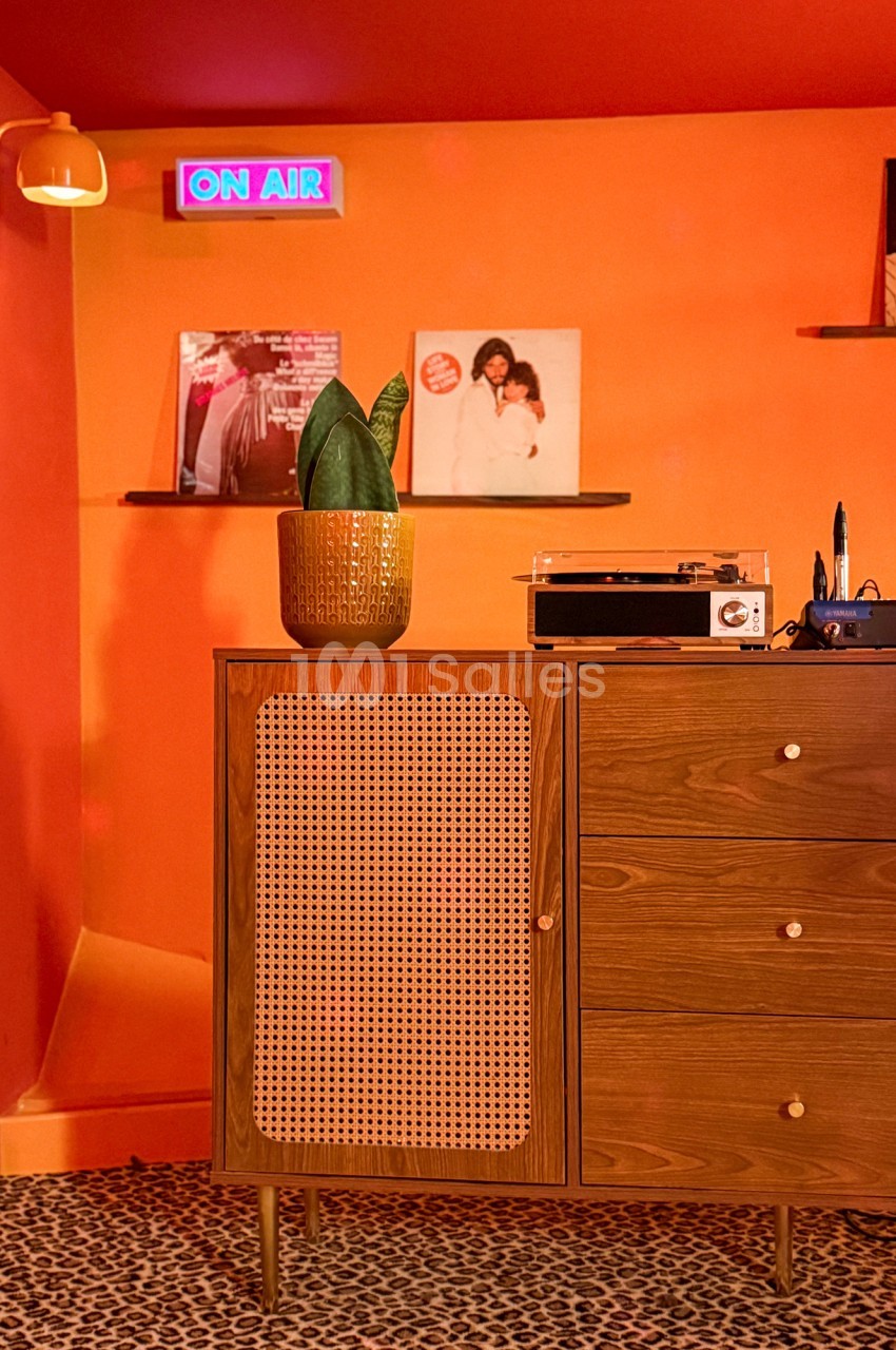 Meuble en bois avec porte en cannage, plante en pot, platine vinyle et disques sur des étagères, mur orange en arrière-plan.