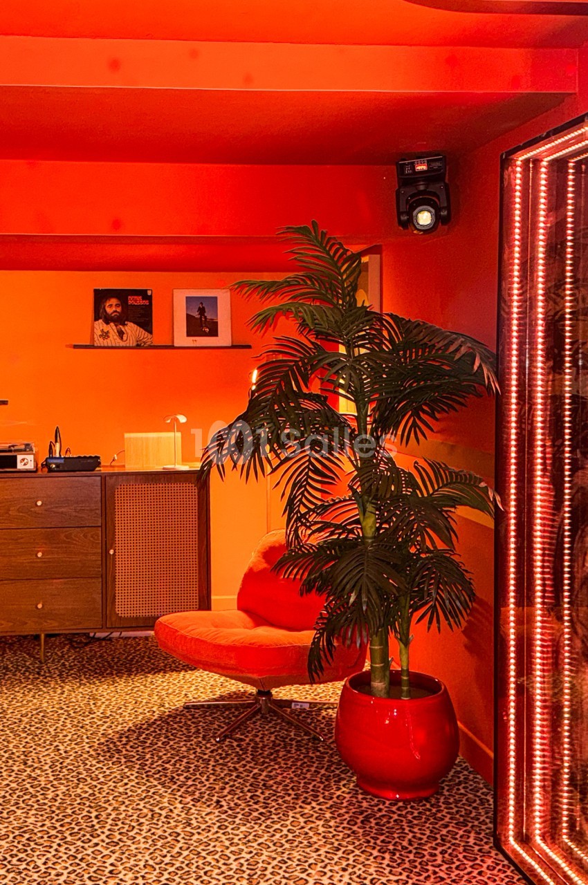 Intérieur rétro avec fauteuil rouge, plante en pot, meuble en bois et murs orange vif, éclairage chaleureux.
