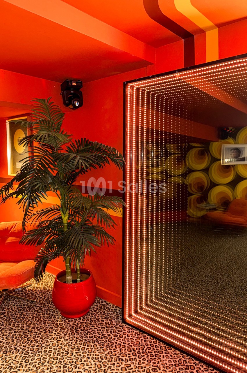 Salle au décor rétro avec murs rouges, fauteuil orange, plante en pot rouge et miroir lumineux à effet infini.
