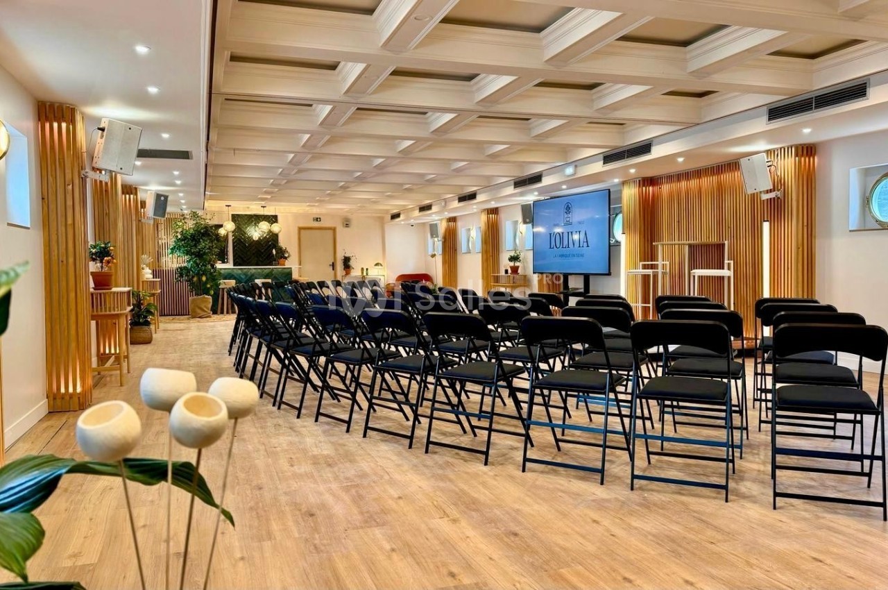 Salle de conférence lumineuse avec des rangées de chaises noires, un écran et une décoration végétale.