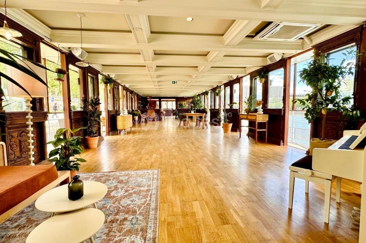 Salle lumineuse avec parquet, grandes fenêtres, plantes décoratives et mobilier en bois clair.