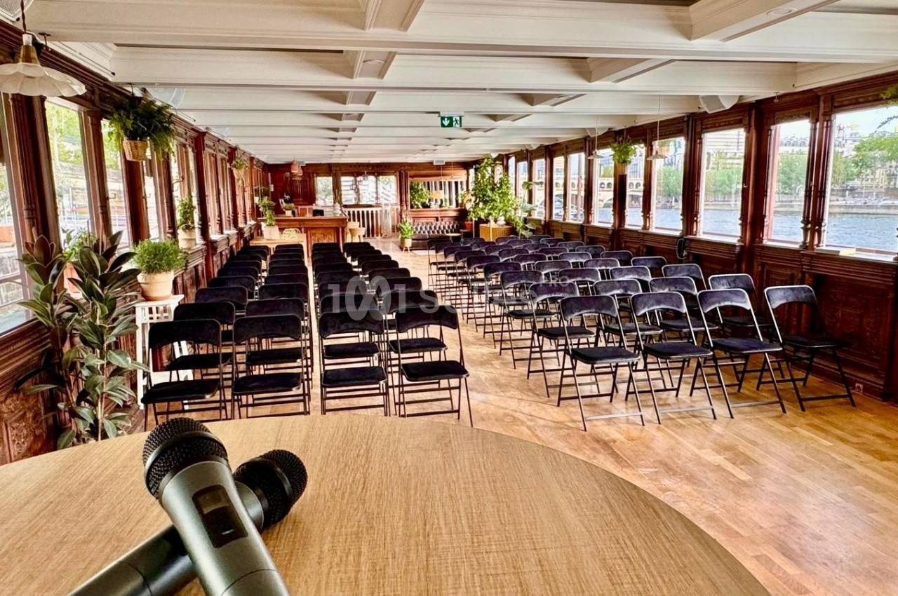 Salle de conférence lumineuse avec des rangées de chaises noires, vue depuis un pupitre avec des micros.