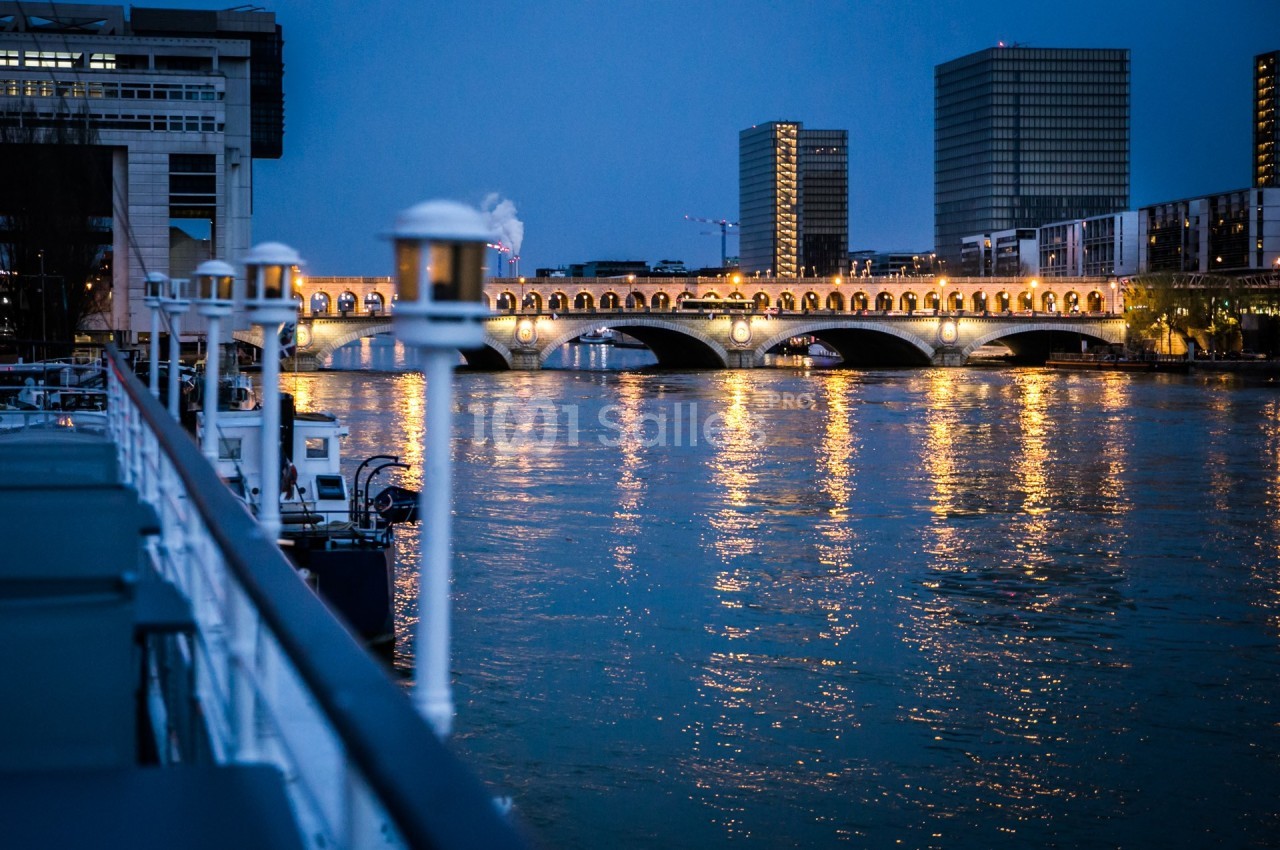Vue nocturne d'un pont illuminé reflété dans une rivière, entouré de bâtiments modernes et de lampadaires.