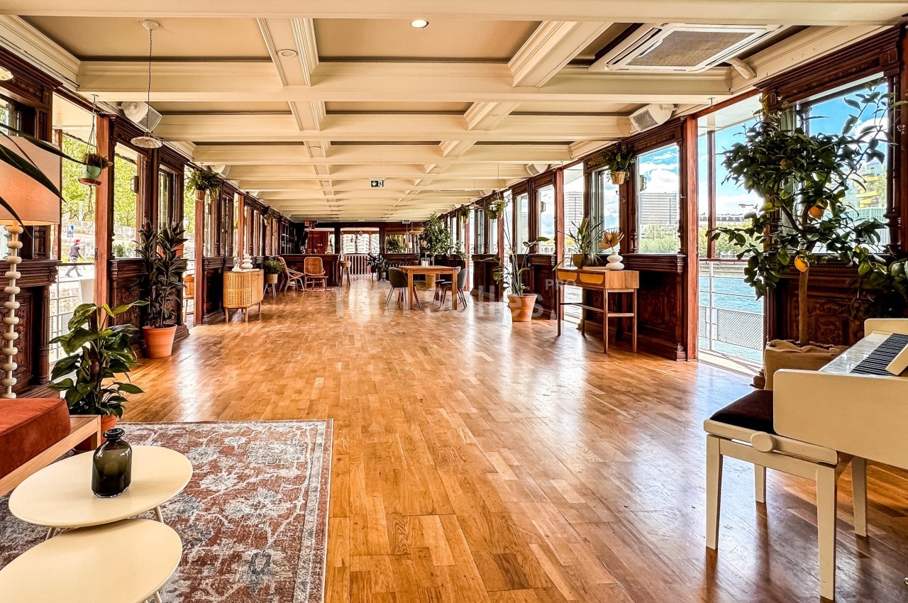 Salle lumineuse avec parquet en bois, grandes fenêtres donnant sur l'extérieur, plantes et mobilier élégant.