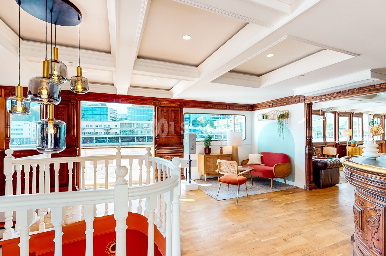 Intérieur lumineux avec escalier blanc, parquet, mobilier moderne et grandes fenêtres donnant sur un paysage urbain.