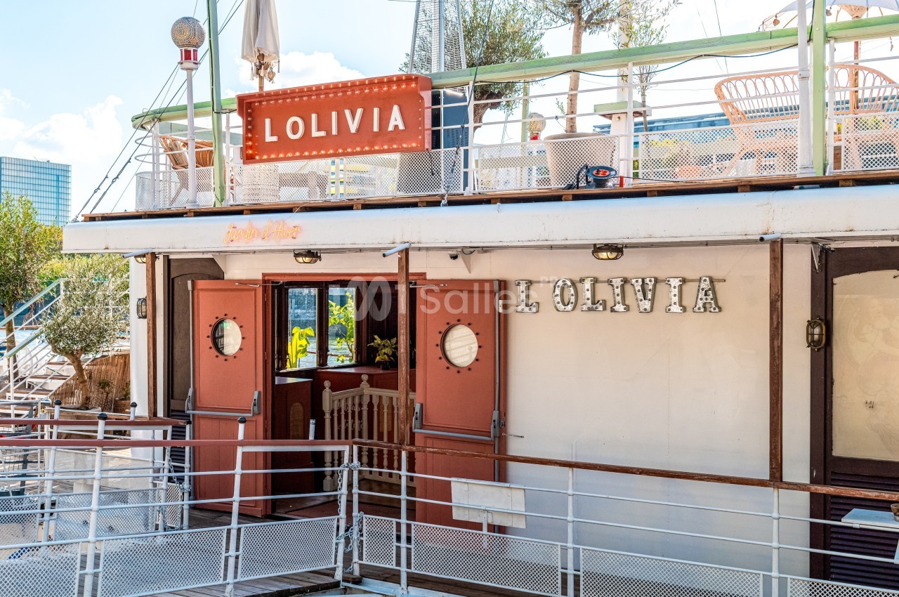 Bateau aménagé avec terrasse, enseigne ’L'Olivia’ visible, portes ouvertes donnant sur l'intérieur.
