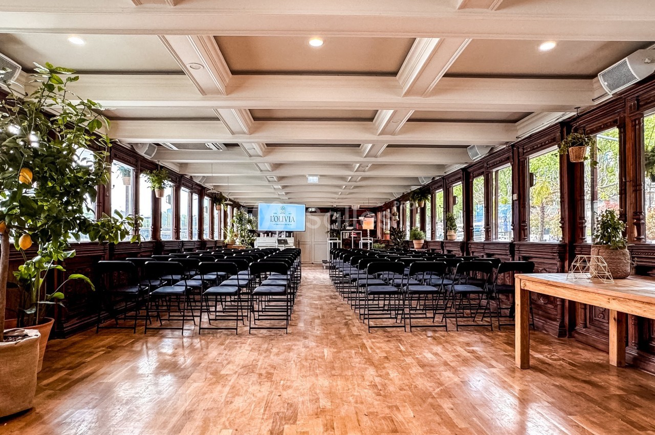 Salle lumineuse avec parquet, rangées de chaises noires, plantes en pot et grandes fenêtres donnant sur l'extérieur.