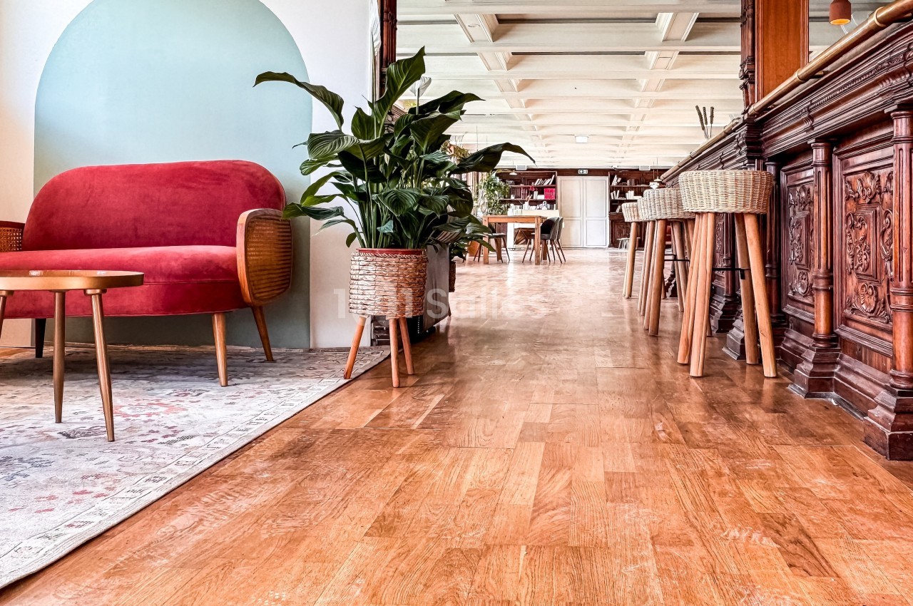 Salon lumineux avec canapé rouge, plantes en pot, mobilier en bois et sol en parquet, menant à un espace ouvert.
