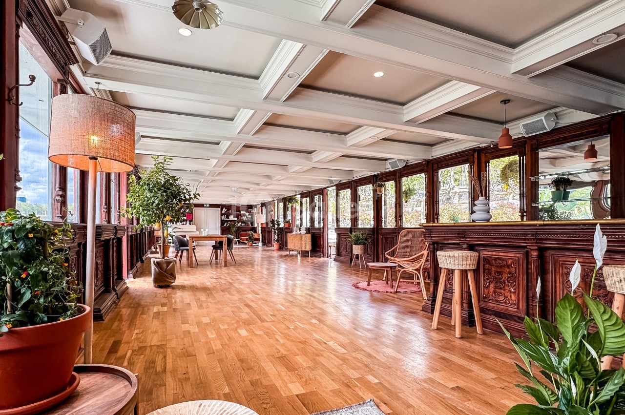 Salle lumineuse avec parquet en bois, grandes fenêtres, plantes en pot et mobilier en bois naturel.