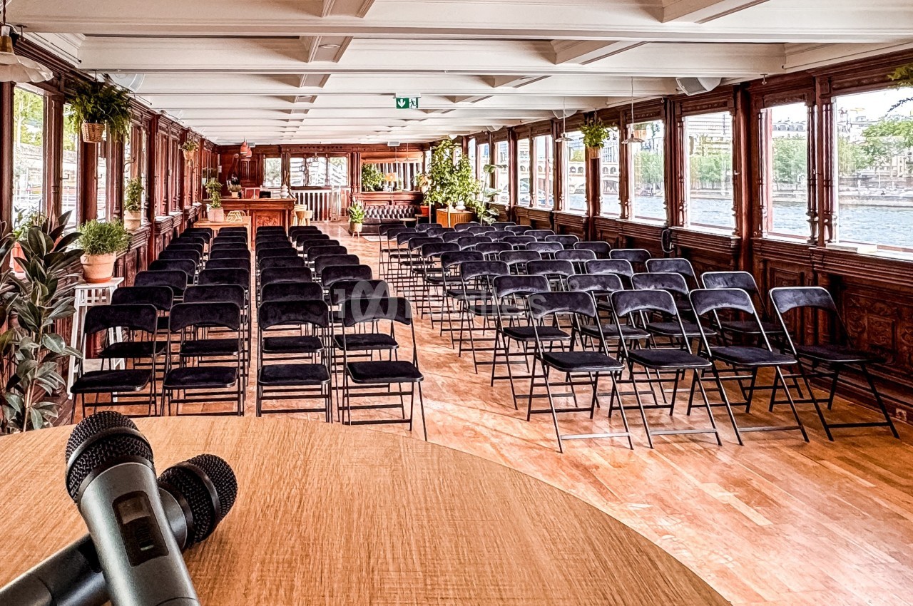 Salle de conférence lumineuse sur une péniche, avec des rangées de chaises noires et des micros sur une table au premier…