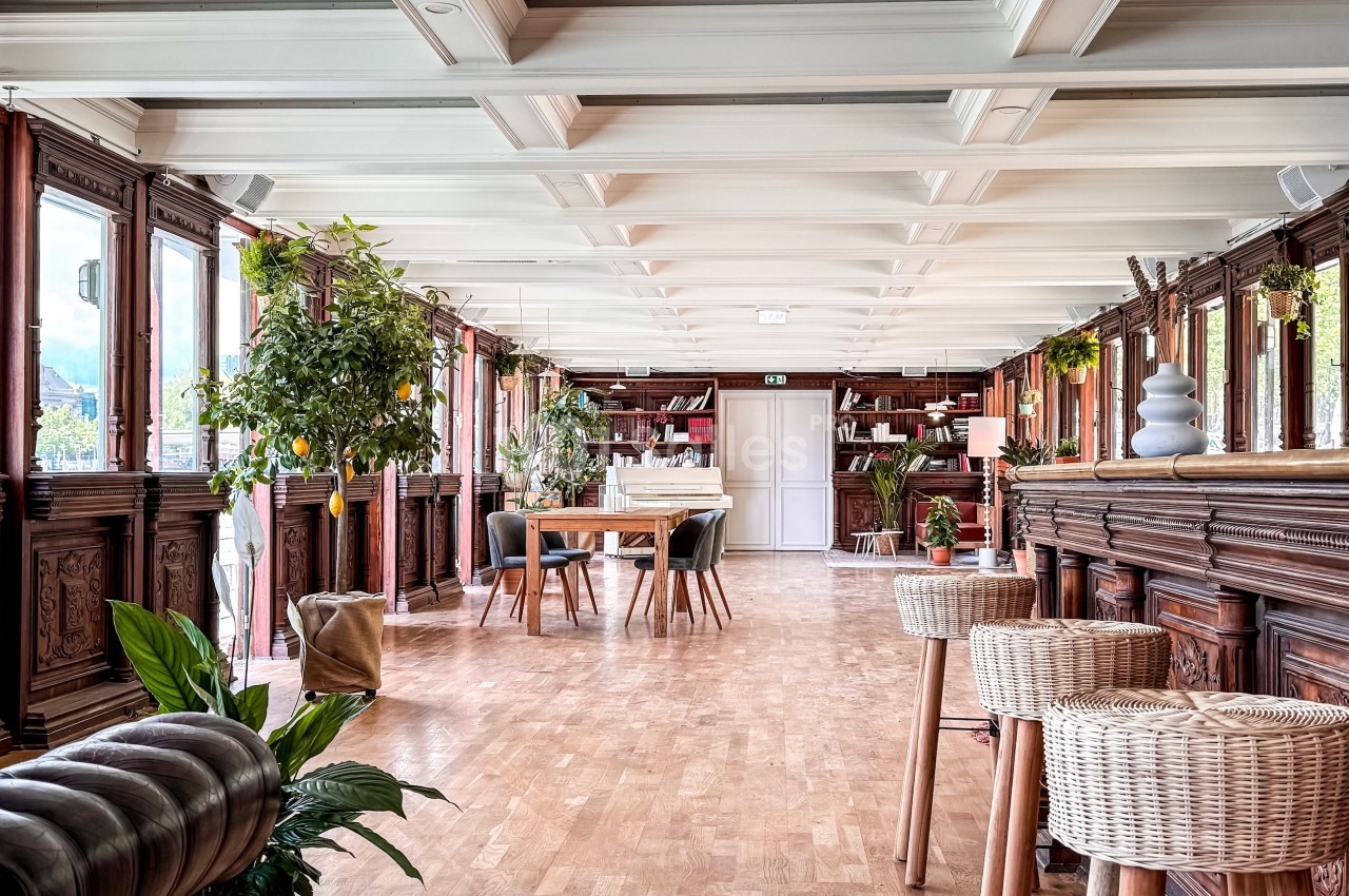 Salle lumineuse avec parquet, mobilier en bois, plantes vertes et grandes fenêtres offrant une vue extérieure.