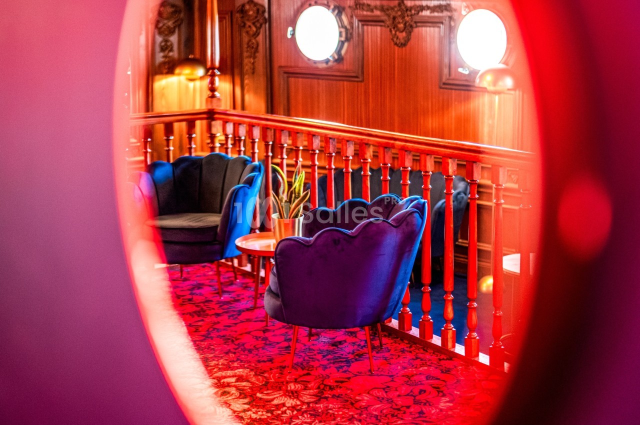 Vue à travers un hublot d'un salon avec fauteuils bleus, table basse et plante, sur un tapis rouge décoratif.