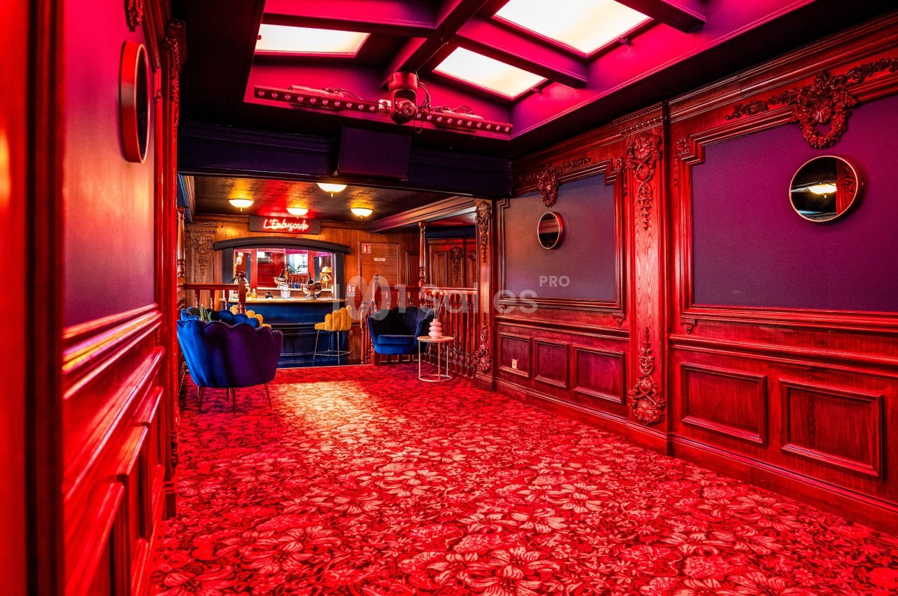 Couloir richement décoré avec moquette rouge, murs en bois sculpté et vue sur un espace lounge éclairé.