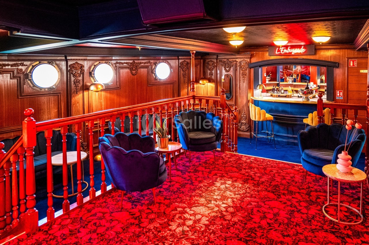 Salon chaleureux avec fauteuils bleus, tapis rouge à motifs, boiseries sculptées et bar éclairé en arrière-plan.