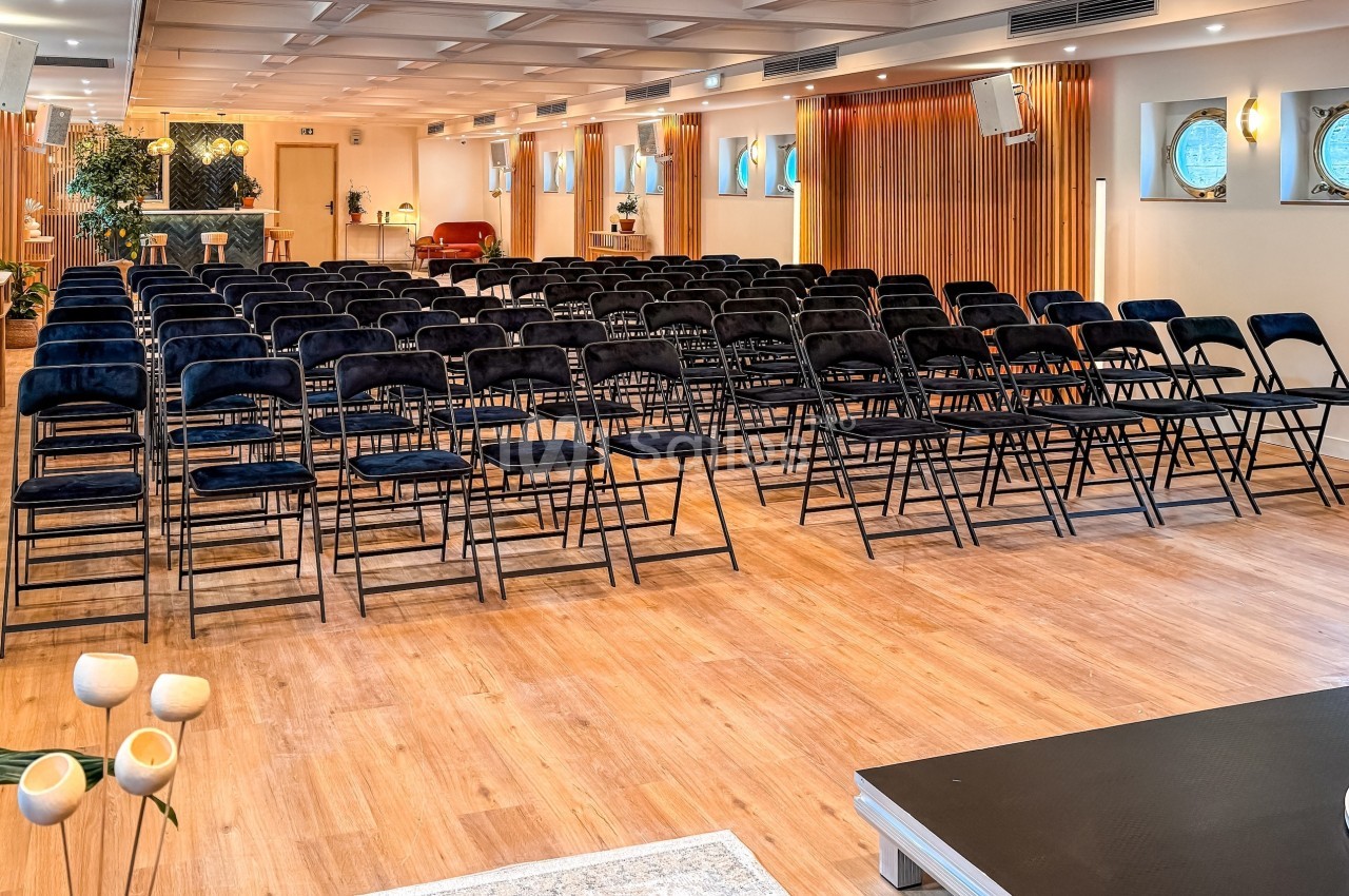 Salle lumineuse avec rangées de chaises noires alignées, parquet en bois clair et décorations modernes.