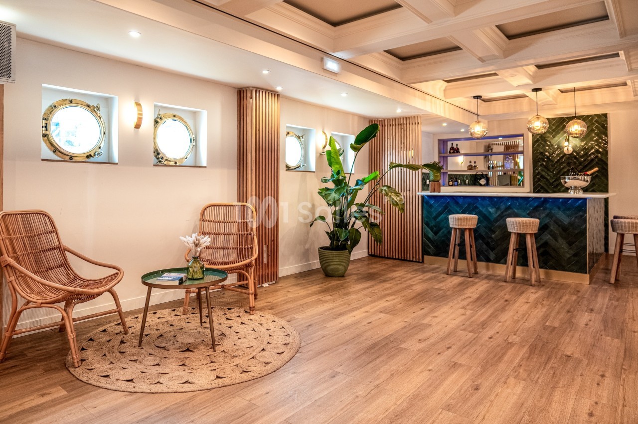 Espace intérieur lumineux avec un bar, des tabourets, des fauteuils en rotin et des plantes décoratives.