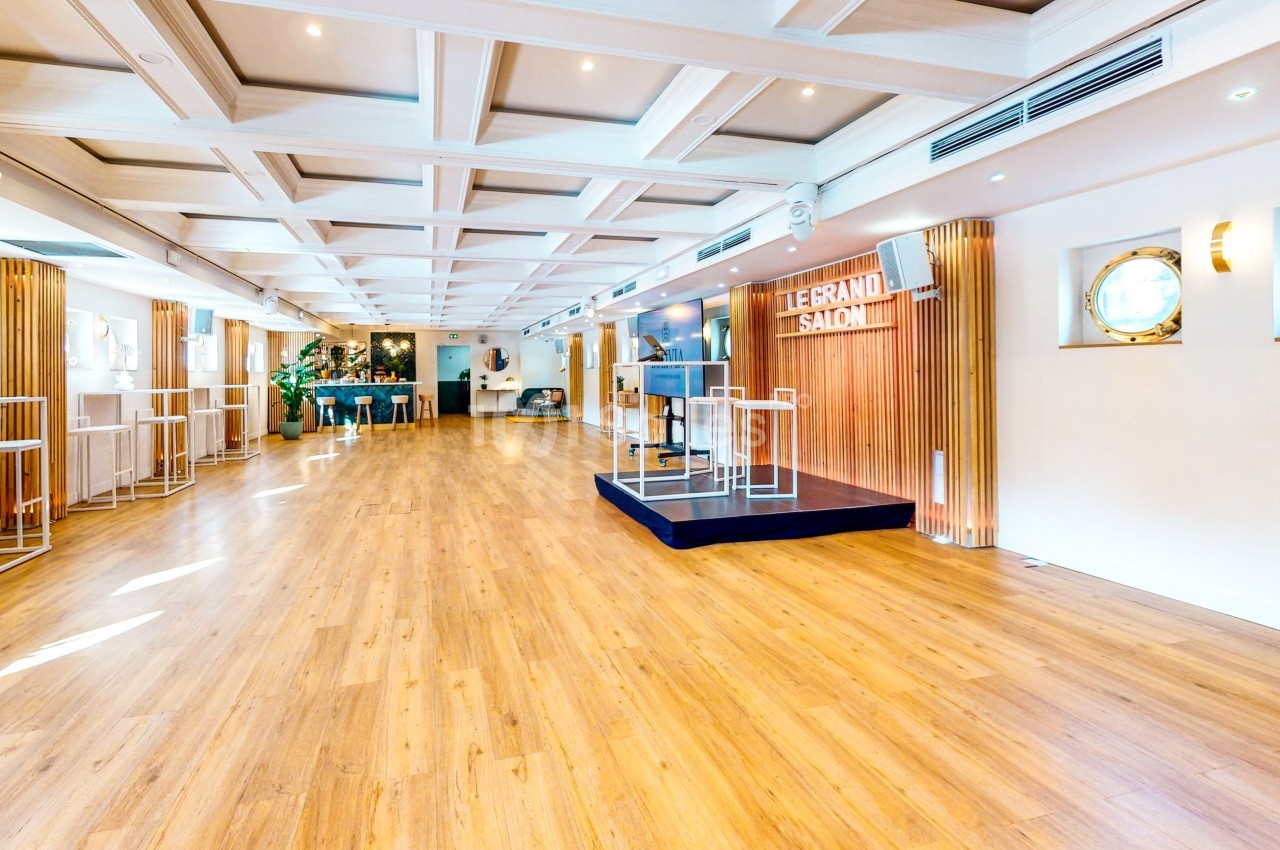 Salle lumineuse avec parquet en bois, murs blancs, hublots ronds et une petite scène surmontée d'un panneau ’Le Grand Salon’.