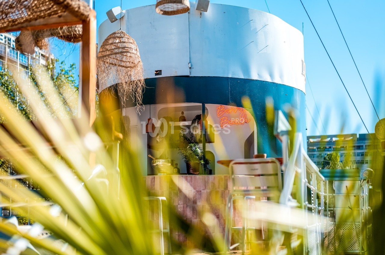 Vue d'un café extérieur avec une décoration estivale, des plantes et des luminaires en osier, partiellement caché par des…