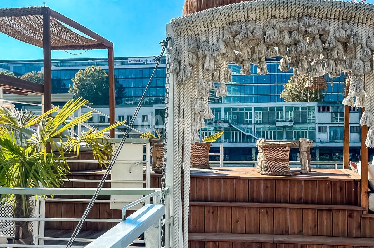 Terrasse en bois avec des plantes et des cordages décoratifs, vue sur des bâtiments modernes en arrière-plan.