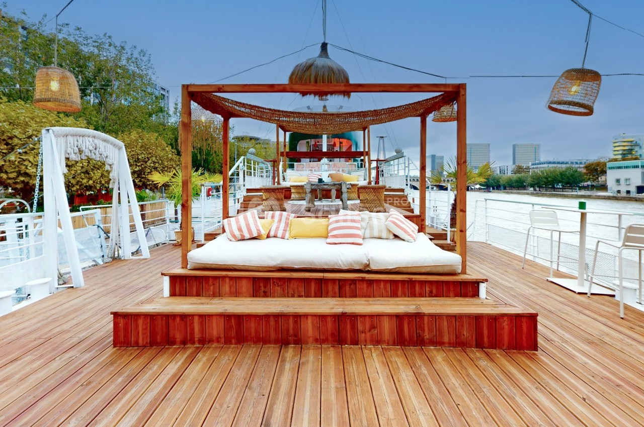 Lit extérieur avec coussins colorés sur une terrasse en bois, entouré de lampes suspendues et vue sur une rivière.