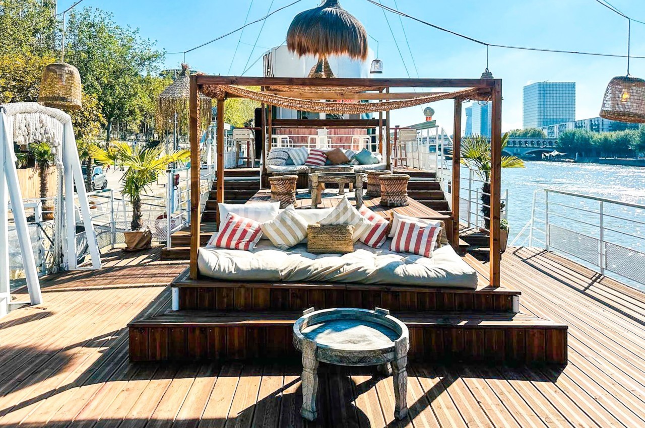Espace détente extérieur avec coussins et lits sous une pergola en bois, situé sur une péniche au bord d'un fleuve.