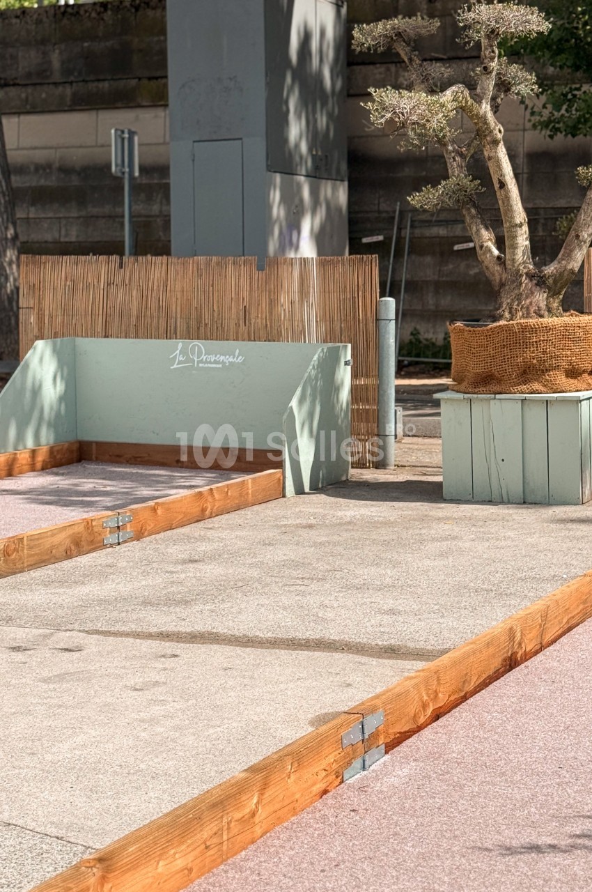 Terrain de pétanque en extérieur avec bordures en bois, un olivier en pot et une clôture en bambou en arrière-plan.