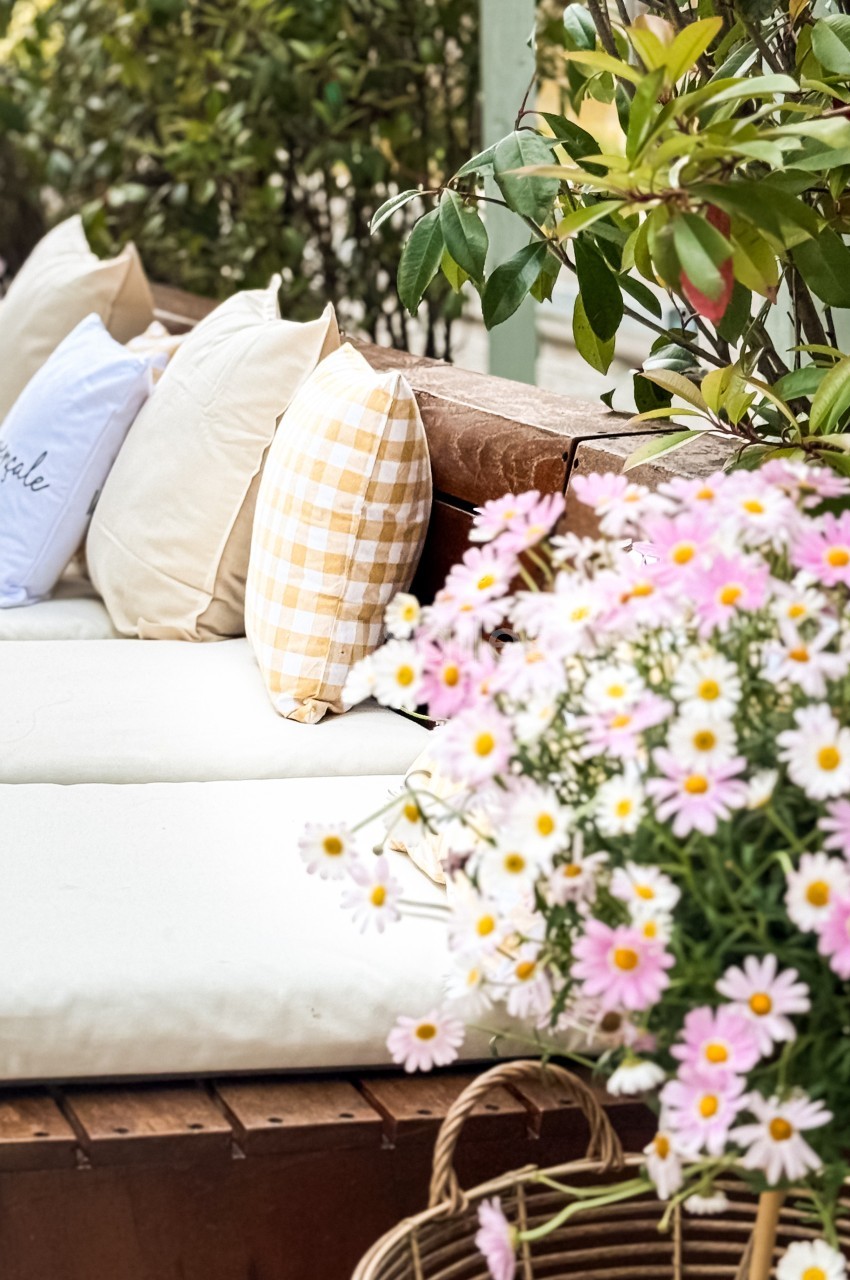 Coussins colorés disposés sur une banquette en bois, entourée de fleurs et de verdure dans un cadre extérieur.
