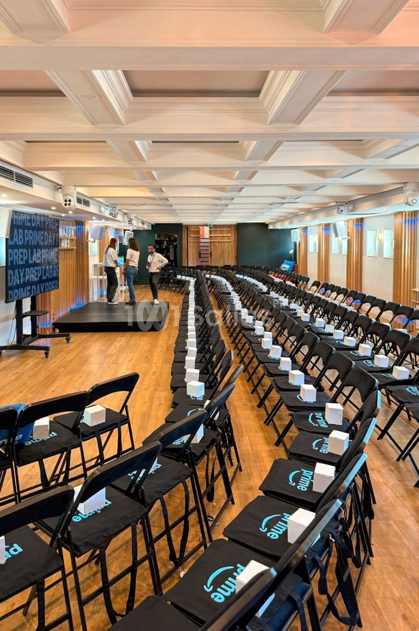 Salle de conférence avec des rangées de chaises noires alignées, une scène et quelques personnes en préparation.
