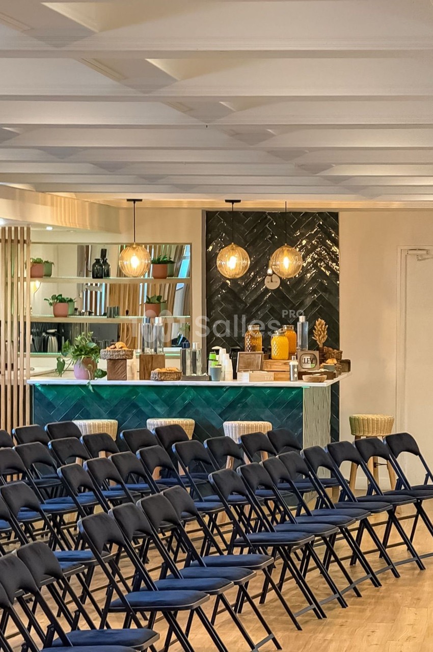Salle de conférence avec des chaises pliantes alignées face à un comptoir de bar décoré et éclairé par des suspensions.