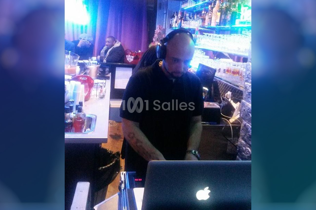 Un DJ portant un casque mixe de la musique sur un ordinateur portable dans un bar éclairé par des lumières bleues.