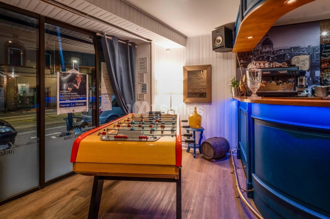 Baby-foot en bois dans un espace intérieur lumineux avec un bar à droite et une grande baie vitrée donnant sur la rue.