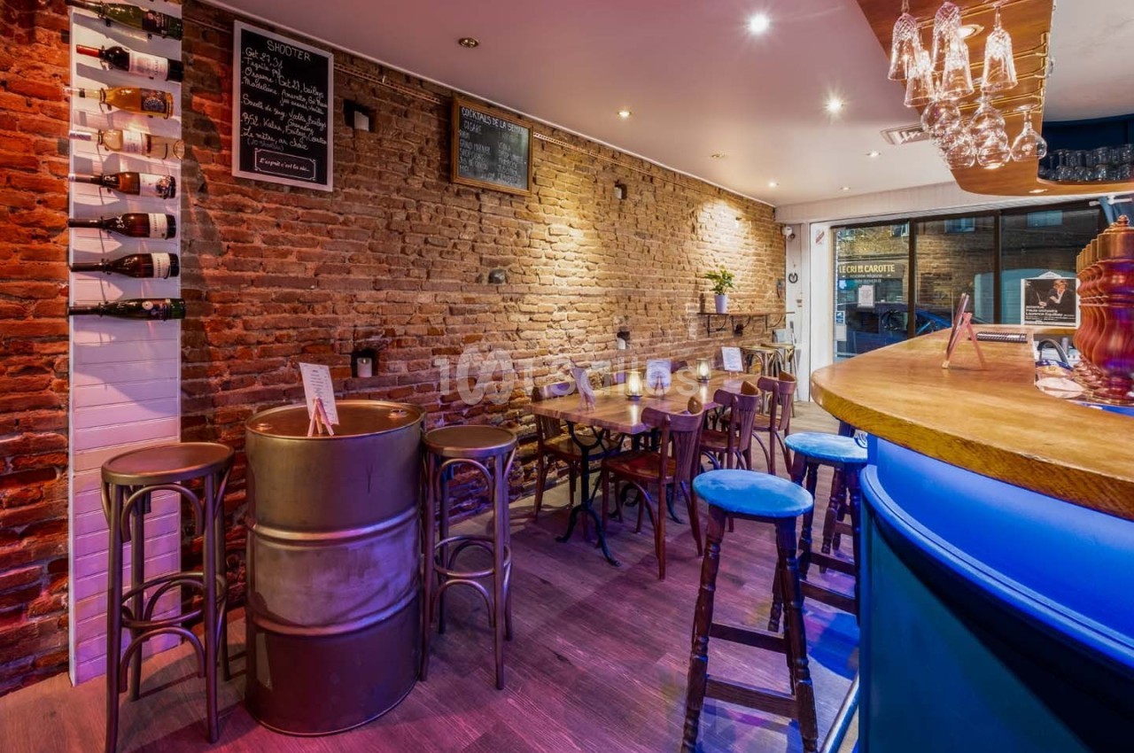 Intérieur d'un bar avec murs en briques, tables en bois, tabourets et comptoir éclairé dans une ambiance chaleureuse.